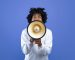 Emotional black teenager making announcement with megaphone on blue studio background. Cheerful African American youth using loudspeaker for sharing news or information