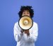 Emotional black teenager making announcement with megaphone on blue studio background. Cheerful African American youth using loudspeaker for sharing news or information